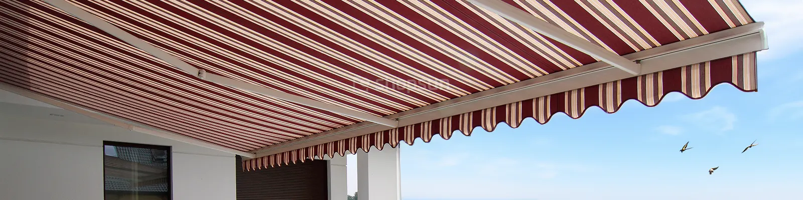 Large patio awning with three arms and fabric in burgundy and beige stripes against a blue sky with three swallows in the background